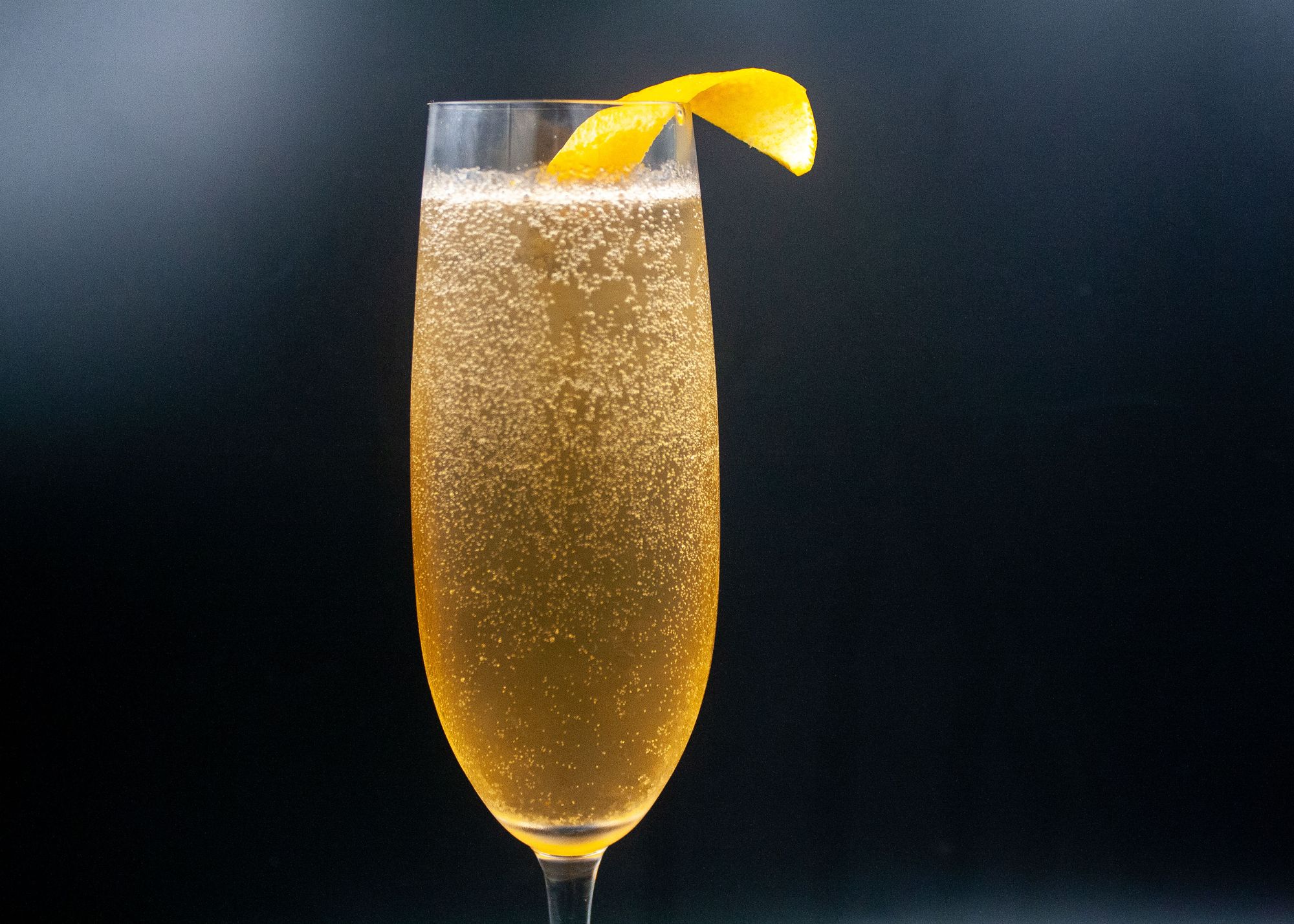 A tall flute filled with a golden bubbly cocktail and garnished with a twist of orange.