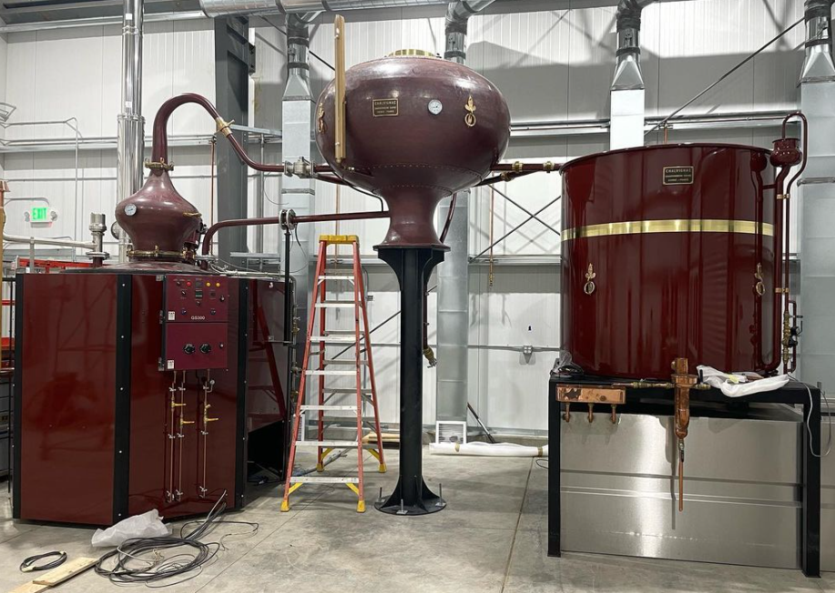 A copper-colored French brandy still showing the elevated wine heating tank flanked by a boiler and condenser.
