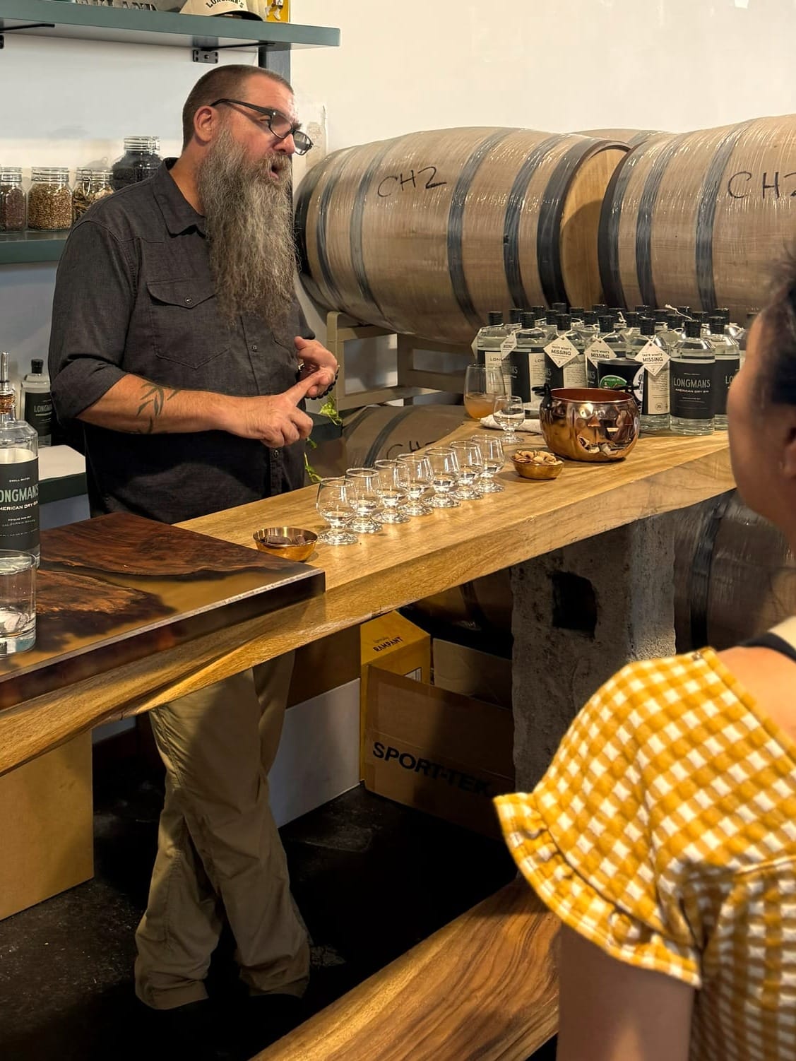 Adam Lamoreaux, head distiller for P&W Spirits Company stands behind a natural wood bar and presents Longman's American Dry Gin to a customer.