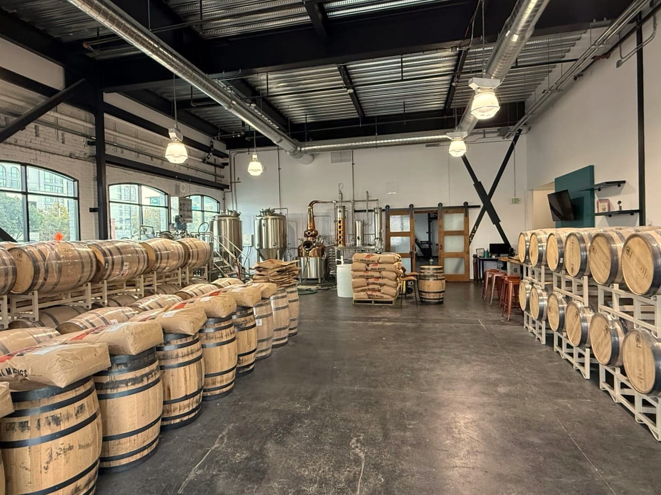A wide photo of a craft distillery interior with a copper and stainless steel still in the background and whiskey barrels stacked with bags of grain in the foreground. 