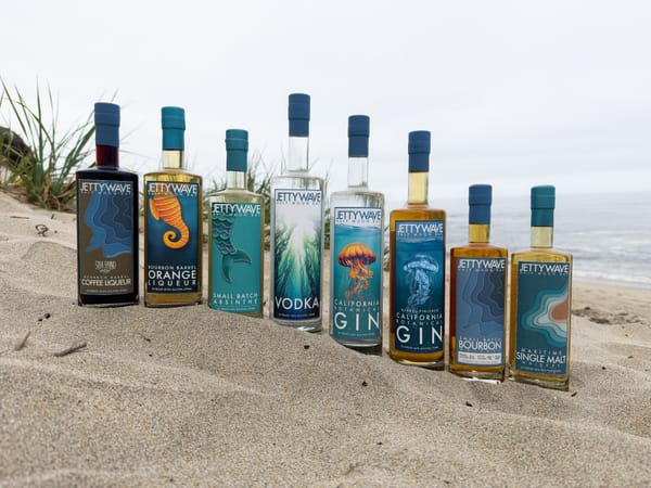 8 bottles of Jettywave liqueurs, gin, vodka, and whiskey in front of a sandy California coastal beach scene