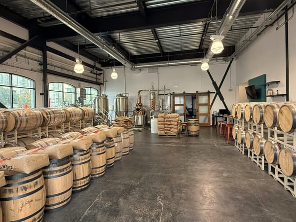 A wide photo of a craft distillery interior with a copper and stainless steel still in the background and whiskey barrels stacked with bags of grain in the foreground. 